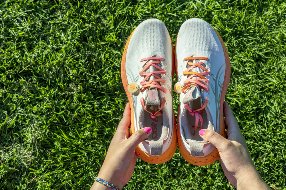 Pair of ASICS x FP Movement GEL-Nimbus 28 running shoes seen from above on grass with coral laces and cream sun charms
